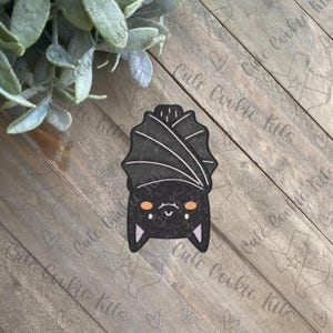 Puede incluir: Un murciélago de dibujos animados negro y blanco con ojos naranjas colgando boca abajo. El murciélago tiene una expresión linda y de dibujos animados.