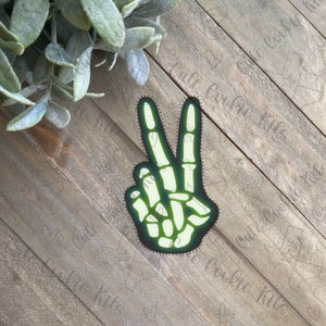 Puede incluir: Un signo de paz verde con un diseño de mano de esqueleto. El signo de paz está delineado en negro.