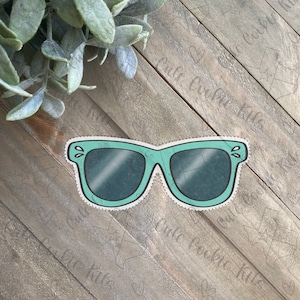 Puede incluir: Gafas de sol azul turquesa con un contorno blanco y un borde punteado. Las gafas tienen un estilo retro con una forma redondeada.