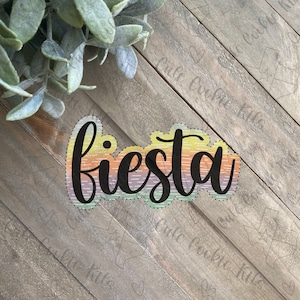 Pode incluir: Um cortador de biscoitos com a forma de "fiesta" em cores de arco-íris com a palavra "fiesta" em letras pretas. O cortador de biscoitos está sobre uma superfície de madeira.