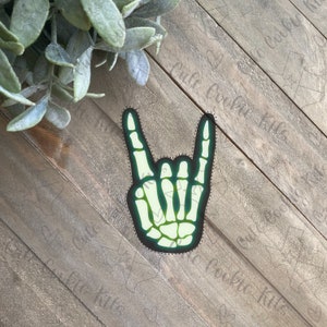 May include: A green and white skeleton hand in the rock and roll gesture. The hand is outlined in black and has a green glow. The image is a cookie cutter.