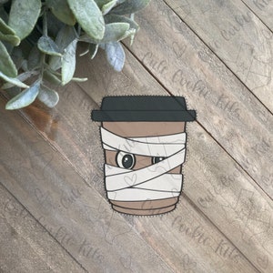 Puede incluir: Una taza de café marrón y blanca con un disfraz de momia. La taza tiene una tapa negra y se ve un ojo.