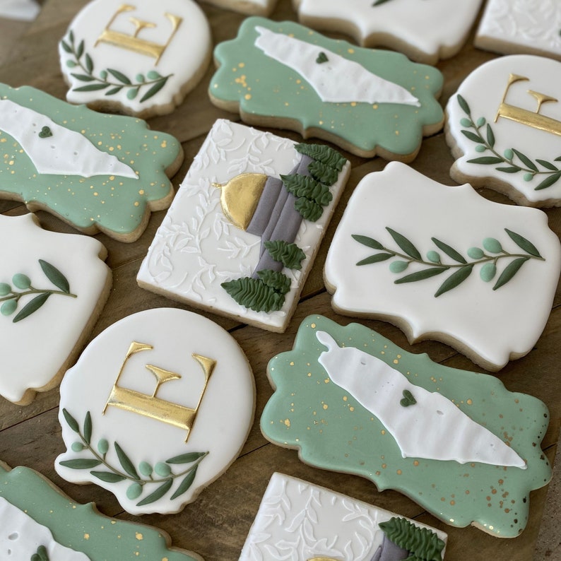 May include: Assortment of decorated sugar cookies with green and gold icing. Some cookies feature the letter "E", while others have a map of Israel with a heart in the center.