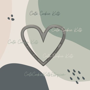 May include: A silver heart-shaped cookie cutter with the text "Cute Cookie Kits" around the edge. The cutter is on a background of green and brown.