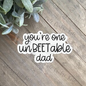 May include: A white sticker with black text that reads "you're one unBEETable dad" on a wood background.