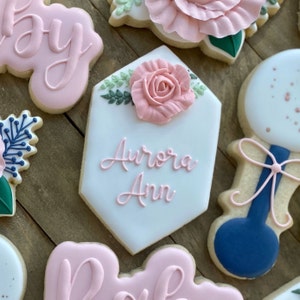 May include: A set of six decorated sugar cookies with pink and blue frosting. The cookies feature a flower, a rattle, and the name "Aurora Ann".