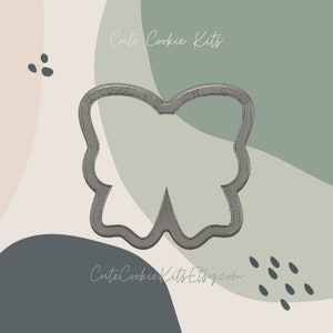 Puede incluir: Un cortador de galletas de metal plateado con forma de mariposa. El cortador está sobre un fondo de formas abstractas en tonos verdes, marrones y beige.