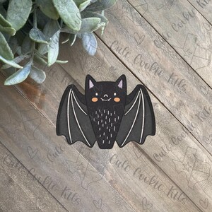 May include: A black bat cookie cutter with a smiling face and small, rounded ears. The wings are spread out and have a textured pattern.