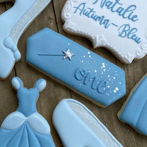 May include: A set of blue and white decorated sugar cookies with a Cinderella theme. The cookies feature a glass slipper, a dress, a wand, and a banner that says "Natalie Autumn-Bleu one".