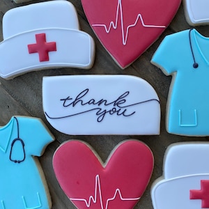 Pode incluir: Conjunto de bolachas de açúcar decoradas com um coração vermelho com uma linha de ECG branca, um chapéu de enfermeira branco com uma cruz vermelha, um top de uniforme azul com um estetoscopio e uma bolacha branca com o texto "obrigado" em preto.