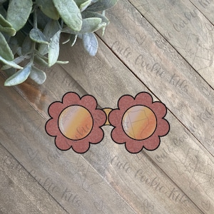 May include: A pair of red flower-shaped sunglasses with orange lenses. The sunglasses are on a wooden background.