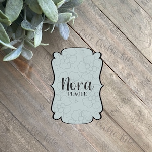 May include: A light blue plaque with a floral design and the name "Nora" in black text.