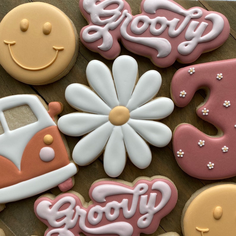 May include: A set of seven decorated sugar cookies. The cookies are decorated with a white daisy, a pink and white groovy sign, a pink number three with white dots, a yellow smiley face, a pink and white groovy sign, a yellow smiley face, and a brown and white van.