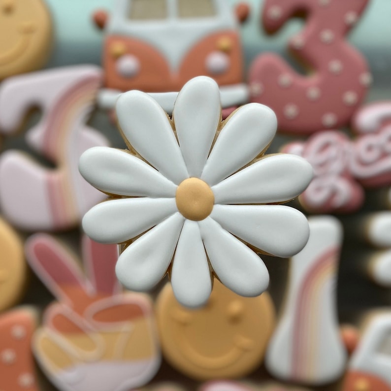 May include: A white daisy cookie with a yellow center. The cookie is surrounded by other cookies, including a peace sign, a rainbow, and a smiley face.
