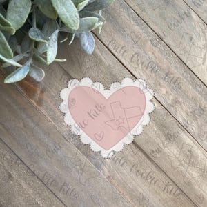 May include: A pink heart-shaped cookie cutter with a scalloped edge. The cutter features a Texas outline with a star and the text "Cute Cookie Kits". A floral design and hearts are also visible.