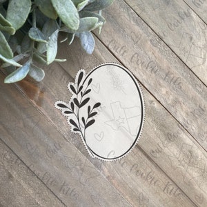 May include: A black and white cookie cutter in the shape of an oval with a floral design and the outline of the state of Texas inside. The cookie cutter is on a wooden surface.