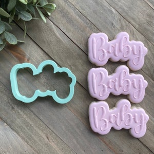 May include: Three pink sugar cookies with the word "Baby" written in cursive on each cookie. A light blue plastic cookie cutter in the shape of the word "Baby" is also shown.