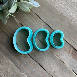 May include: Three blue plastic cookie cutters shaped like kidney beans. The cutters are different sizes and have the words "Cute Cookies" and "Made with Love" on them.