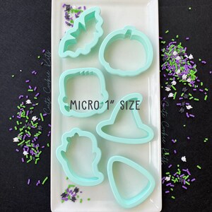 May include: Set of six light blue plastic cookie cutters in various Halloween shapes: a bat, pumpkin, ghost, witch hat, Frankenstein, and a candy corn. The cutters are arranged on a white surface with the text "MICRO 1" SIZE" printed in black.