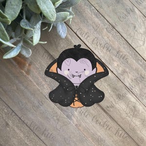 Puede incluir: Un cortador de galletas de vampiro de dibujos animados morado y negro con una capa y colmillos.