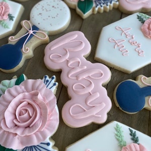 May include: A set of decorated sugar cookies with a pink and blue colour scheme. The cookies feature a floral design, a baby bottle, the word "Baby" in pink script, and a hexagon-shaped cookie with the text "Aurora Ann".