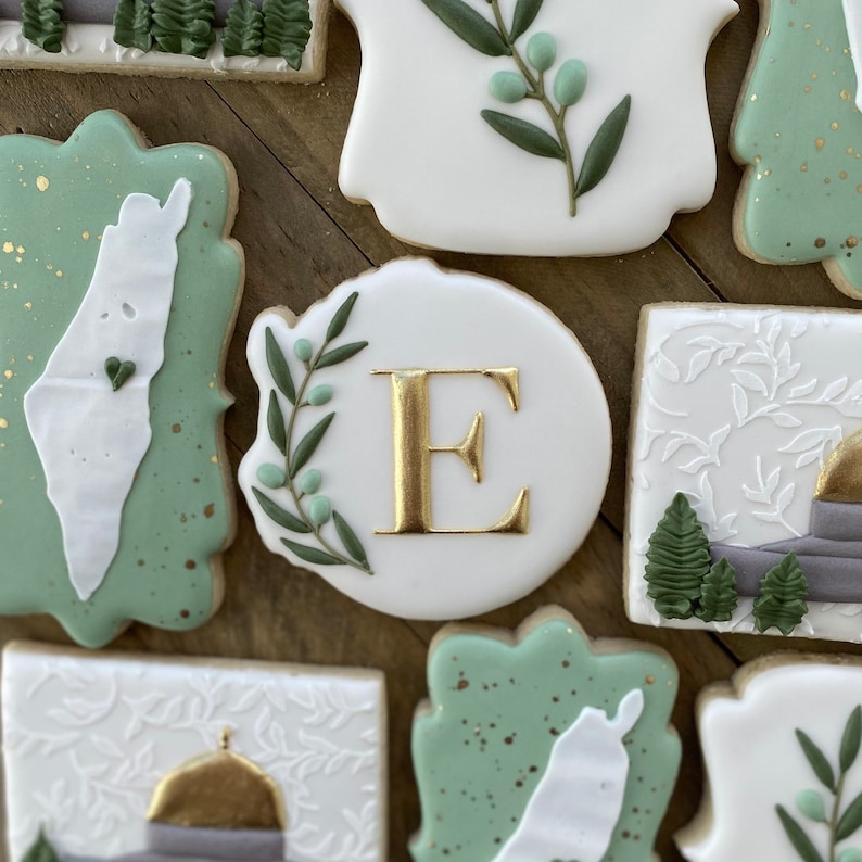 May include: Assortment of decorated sugar cookies with green and white icing. The cookies feature a map of Israel, a leafy branch, the letter E, and a building with a golden dome.