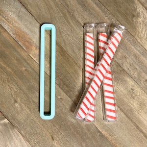 May include: Four red and white striped candy canes in clear plastic packaging. The candy canes are arranged in an X shape. A light blue rectangular cutout is in the background.