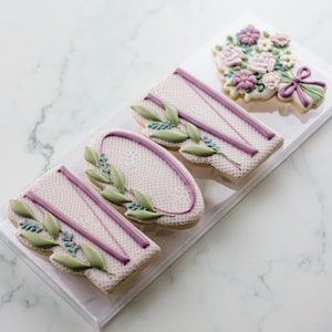 Puede incluir: Cuatro galletas de azúcar decoradas en una caja de plástico transparente. Las galletas tienen forma de las letras "MOM" y un ramo de flores. Las galletas están decoradas con encaje blanco, glaseado morado y hojas verdes.