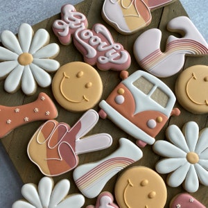 May include: A collection of 12 decorated sugar cookies with various designs, including peace signs, smiley faces, daisies, and a vintage van. The cookies are decorated in shades of brown, orange, pink, and white.