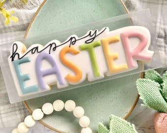 May include: A white cookie decorated with colorful icing spelling "Happy Easter". The cookie is in a clear plastic bag and sitting on a light green plate.