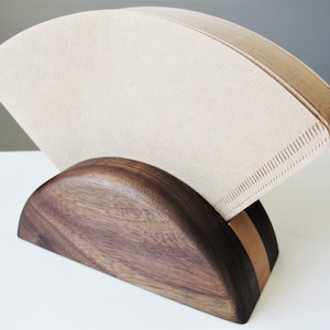 May include: A wooden coffee filter holder in a dark brown finish, holding a stack of beige paper filters. The holder has a curved design with a lighter brown accent stripe.