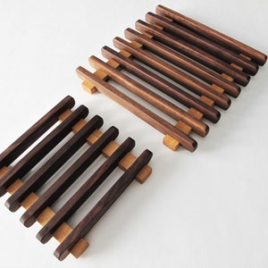 May include: Two wooden trivets with a slatted design. The trivets are made of dark brown wood with lighter brown supports. The trivets are designed to protect surfaces from hot dishes and are a kitchen accessory.