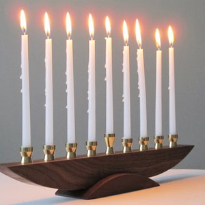 May include: A wooden menorah with nine lit white candles. The candles are set in brass holders on a curved wooden base. The flames are bright and the background is a neutral grey.