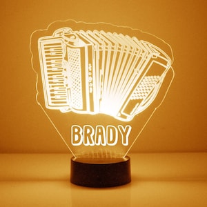 Può includere: Una luce notturna bianca illuminata a forma di fisarmonica con il nome "BRADY" stampato sotto. La luce notturna è su una base nera.