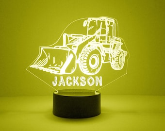 May include: A white outline of a yellow construction vehicle with a scoop, on a black base with the name "JACKSON" underneath. The vehicle is illuminated from behind, casting a yellow glow.