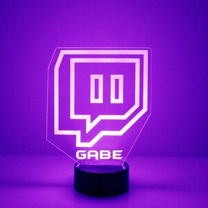 Op de afbeelding: Een paars verlichte acryl bureaulamp met het Twitch-logo, een gestileerde tekstballon met twee verticale rechthoeken. De naam "GABE" is eronder gegraveerd. De lamp staat op een zwarte voet.