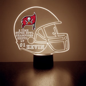 Peut inclure: Contour blanc d'un casque de football américain avec un logo des Buccaneers et le texte "2 TIME SUPER BOWL CHAMPIONS XXXVII LV #1 KEVIN" sur une base noire.