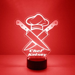 May include: A red illuminated acrylic lamp with a chef's hat and crossed knives design. The lamp has the text "Chef Kelsey" and a black base. The design is a kitchen-themed decorative item, suitable for a kitchen or dining area.