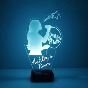 May include: A blue and white night light with a silhouette of a person singing into a microphone. The light has a star and musical notes around it. The text "Ashley's Room" is at the bottom of the light.