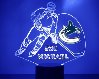 May include: A white 3D printed hockey player with a stick and a Vancouver Canucks logo. The player's number is 20 and the name Michael is printed below the logo. The base of the lamp is black.