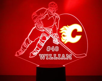 May include: A white 3D printed hockey player with a stick and a flaming C logo. The player is wearing a jersey and pants. The base of the lamp is black. The text on the base reads "#40 WILLIAM".