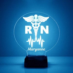 May include: White LED night light with a round, clear acrylic base. The base features a white silhouette of a medical caduceus symbol with the letters "RN" and a heartbeat graphic. The name "Maryanne" is written below the graphic.