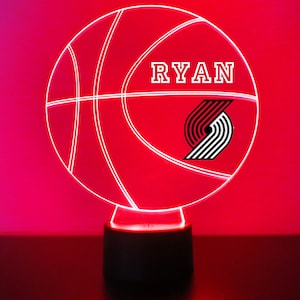 May include: A red basketball-shaped night light with a white outline of a basketball and the word "RYAN" in white letters. The Portland Trail Blazers logo is in the lower right corner of the light.