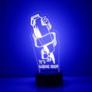 Puede incluir: Un letrero blanco iluminado con un gráfico de unas tijeras de barbero envueltas en una cinta. El letrero dice "Ty's Barber Shop".