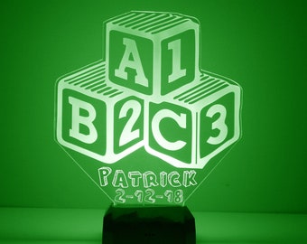 May include: A white acrylic night light shaped like three stacked blocks with the letters A1, B2, and C3 on them. The base of the night light is black and has the name Patrick and the date 2-12-18 engraved on it.
