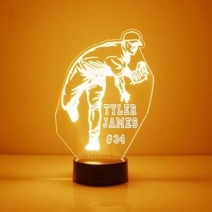 May include: A white illuminated acrylic night light featuring a baseball player in a pitching pose. The light has a black base and includes the name "Tyler James" and the number "#34".