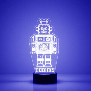以下が含まれることがあります： 黒い台座が付いた白いロボットのナイトライト。ロボットは、目、口、鼻のついた顔をしています。ロボットの下には「TOMMY」という名前が書かれています。
