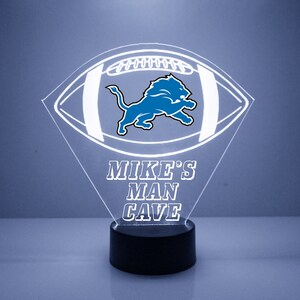 May include: A white illuminated acrylic football with a blue Detroit Lions logo and the text "MIKE'S MAN CAVE" on a black base.
