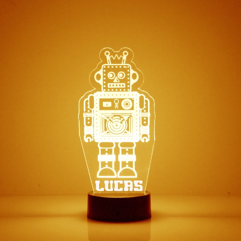 以下が含まれることがあります： ベースに「Lucas」という名前が書かれた白いロボットのナイトライト。ロボットは、2本のアンテナが付いた丸い頭と、中央に丸い窓のある四角い胴体を持っています。ナイトライトは黒いベースの上に置かれています。