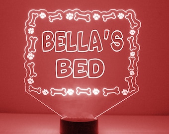 May include: A white LED night light with a clear acrylic base and a white frame. The frame is decorated with paw prints and bone shapes. The light features the text "BELLA'S BED" in white.